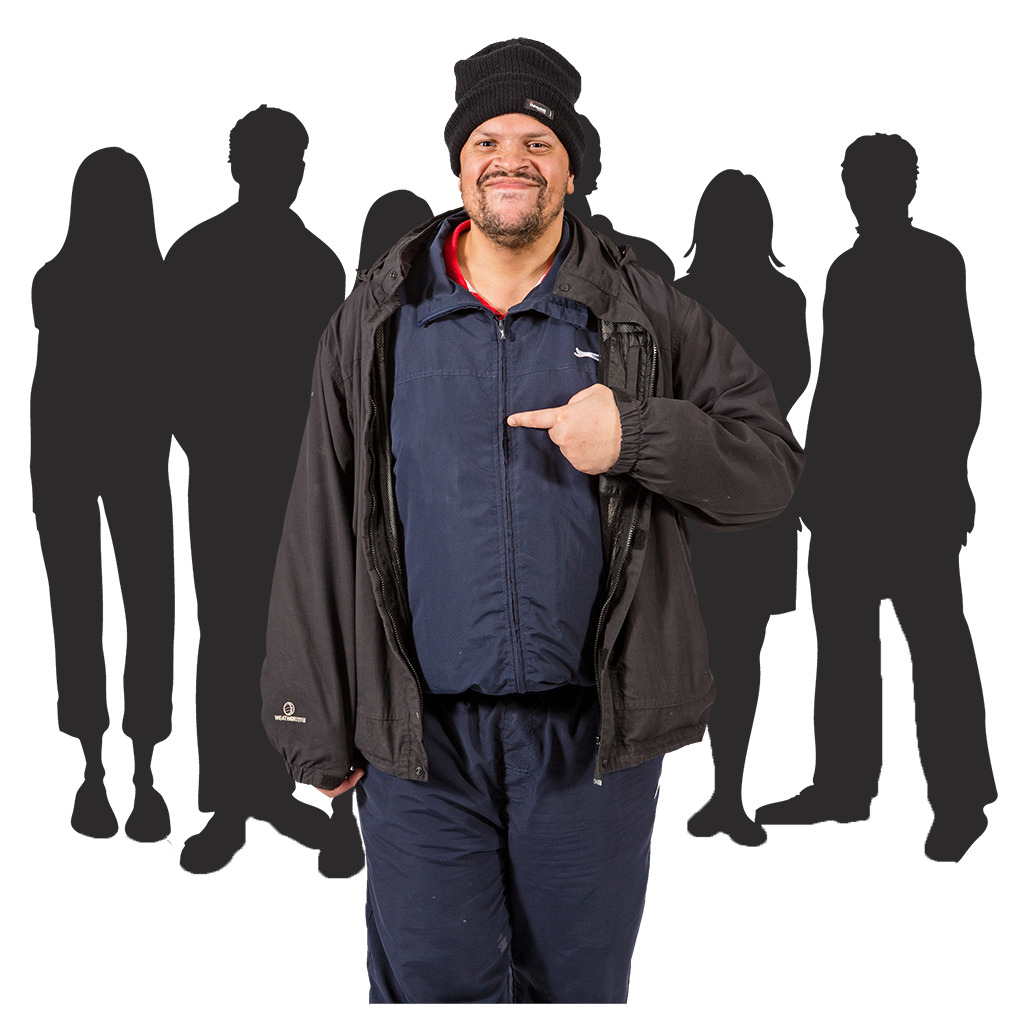 A man smiling and pointing at himself, wearing a black beanie and jacket, with blurred silhouettes of people in the background.