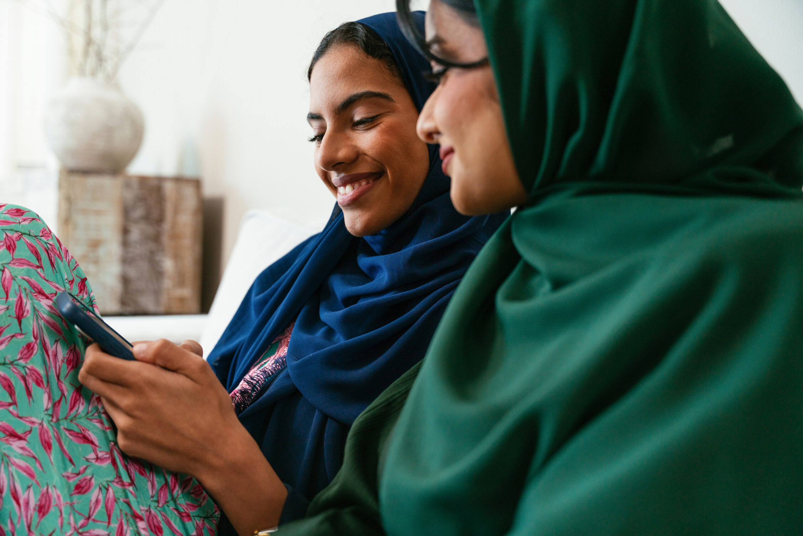 Two emirati women, wearing traditional abayas and hijabs, are sitting together in their living room, enjoying a moment of connection as they look at a smartphone and share a smile