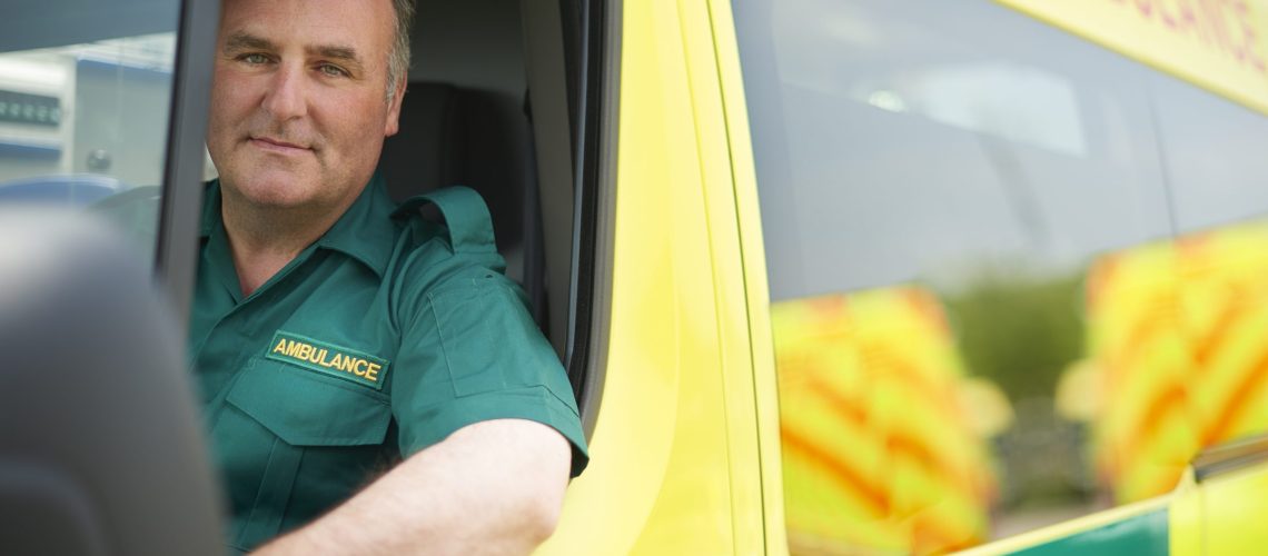 uk ambulance staff members in his ambulance. he is wearing  green ambulance uniform typical of uk paramedics. he is sitting in the ambulance ready to go to a call